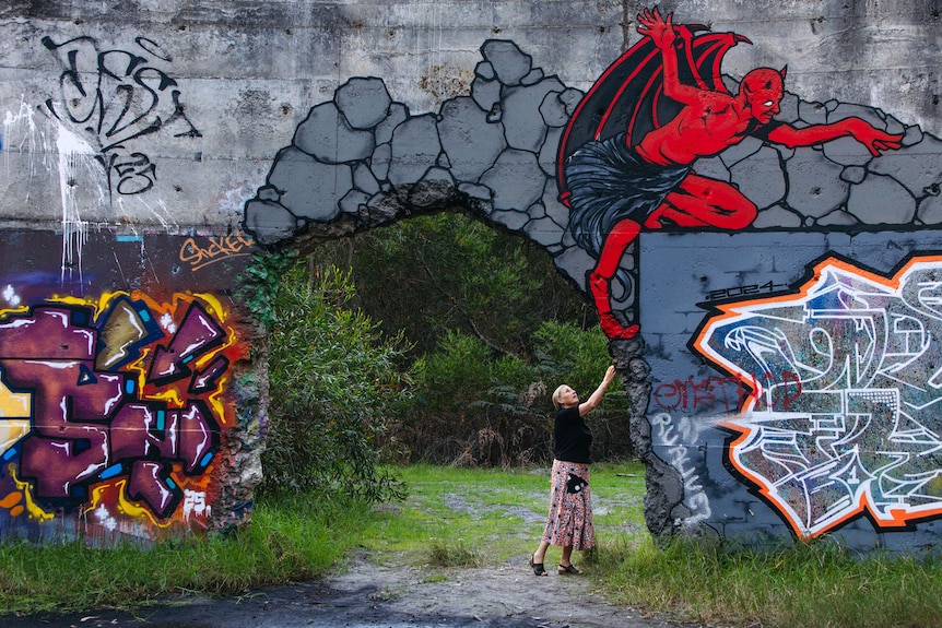 A woman stands beneath a concrete archway in a huge wall covered with graffiti.