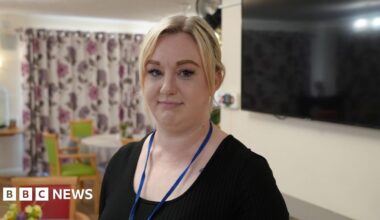 Kerrie Wollen standing in a dining room in a care home. She has blonde hair and is wearing a black T-shirt. Behind her is a flat-screen TV on a wall and tables laid for a meal.