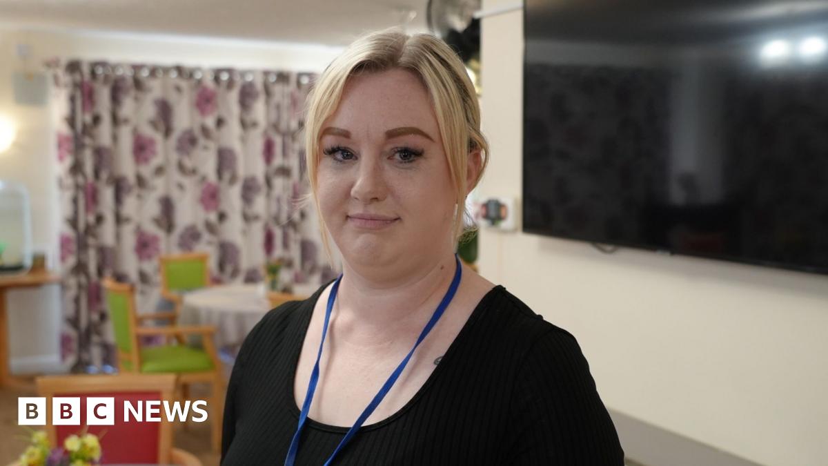Kerrie Wollen standing in a dining room in a care home. She has blonde hair and is wearing a black T-shirt. Behind her is a flat-screen TV on a wall and tables laid for a meal.
