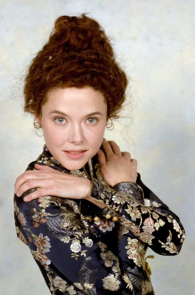Annette Bening in a navy dress with metallic adornments crosses her arms and holds her hands on her shoulders.