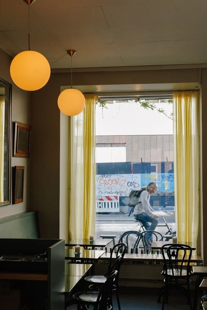 The interior of a bistro-style wine bar, with mint green interiors, sheeny furniture, and wooden chairs, and a window in front of which a cyclist is passing by.