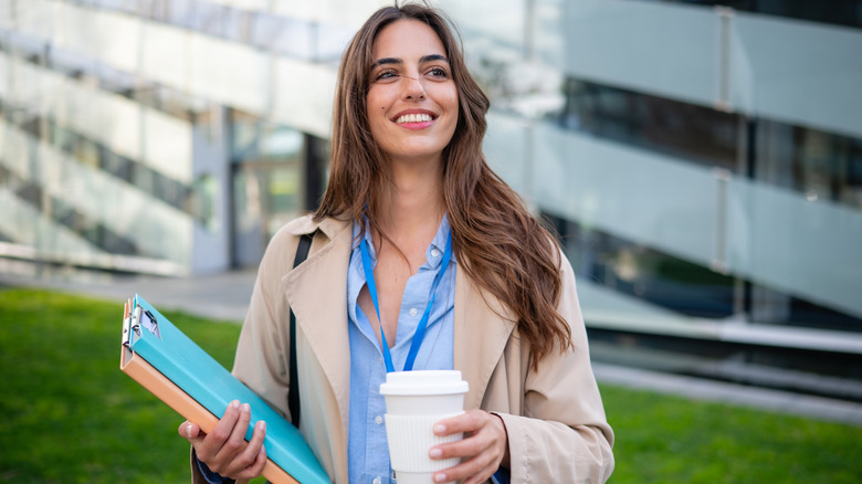 A woman standing outdoors holding a folder and a coffee, wearing a lanyard around her neck