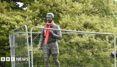 Fenced off statue of Brian Clough wearing a red scarf. There are trees in the background.