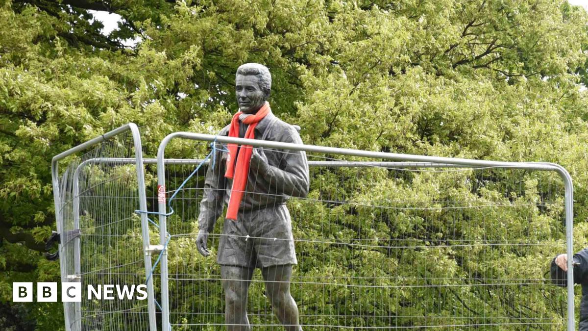 Fenced off statue of Brian Clough wearing a red scarf. There are trees in the background.
