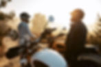 Two riders stand next to a yellow and white motorcycle in the foreground. Warm summer tones and a sandy base.