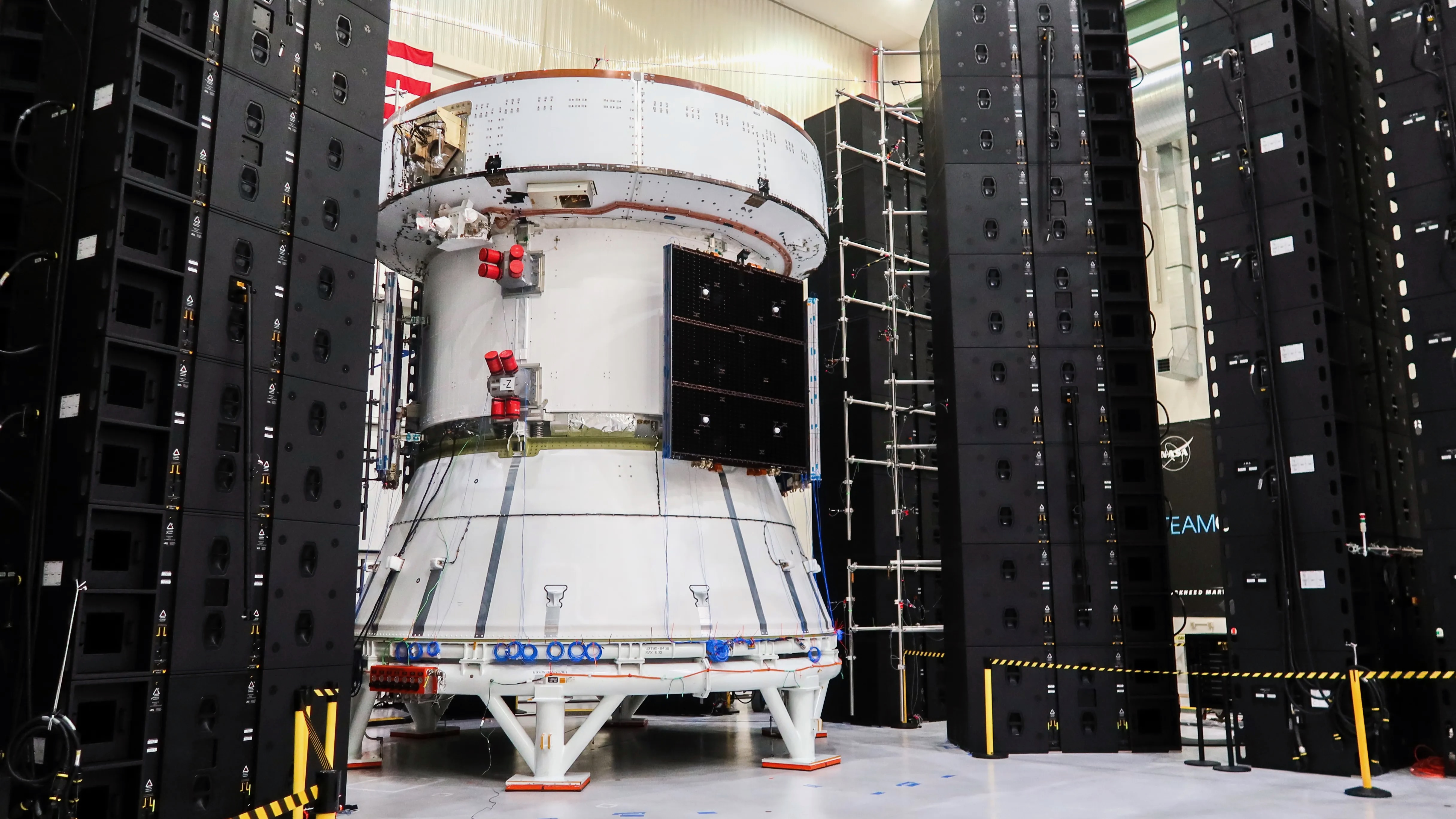 a white space capsule sits in a big, white-walled room, surrounded by black scaffolding