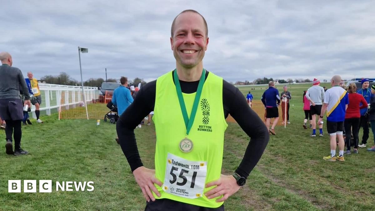 Wood wearing a yellow running vest, with a running track and other runners in the background, resting his hands on his hips, smiling into the camera. He is wearing a medal on a green strap around his neck.