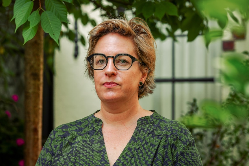 Angela with short hair wearing glasses and a green print top standing in front of a building with shrubs around