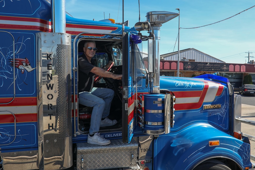 A man in jeans, a black polo and sunglasses smiles from inside the cab of his blue and red truck.