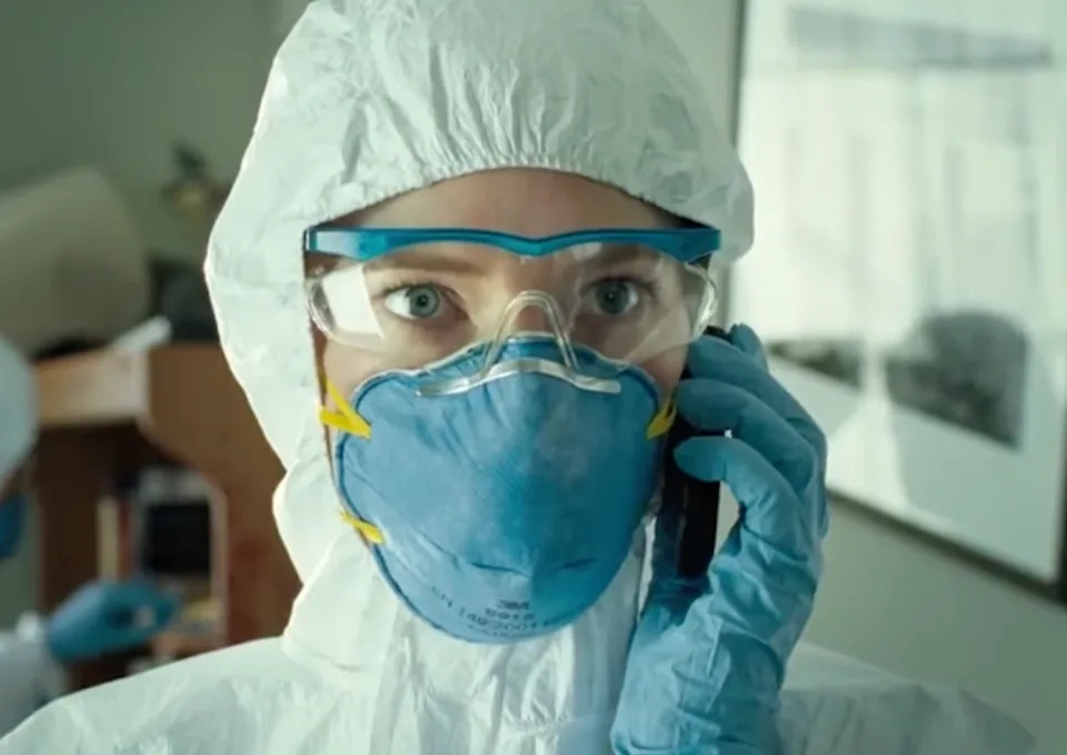 Person in protective gear, including goggles and a mask, holding a phone to their ear in a medical or lab environment