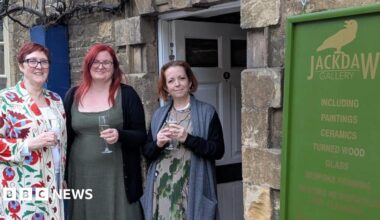 Three women standing outside a stone doorway beneath a green ‘Jackdaw Gallery’ sign, each holding a drink; the gallery entrance and signage are visible behind them.