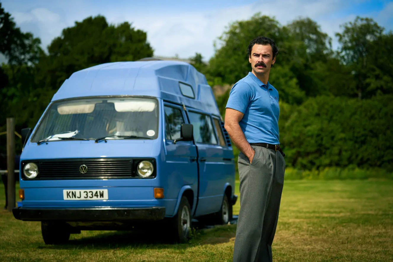 A man in a blue polo shirt and gray trousers stands in a grassy field next to a blue camper van.