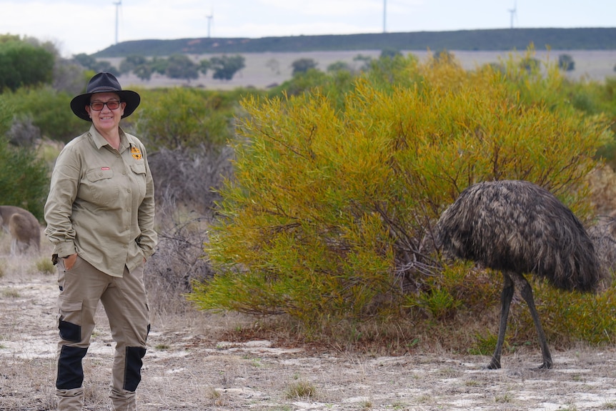 Middle aged woman stands in a bushy paddock next to an emu and kangaroo.