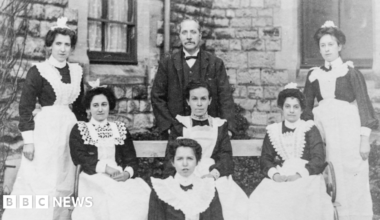 Three rows of women wearing Victorian hospital uniforms - dark dresses and white aprons - and a man are pictured sitting or standing in front of a stone building.