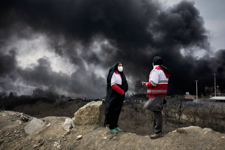 two women in hijabs and red and white jackets wearing face masks, large plumes of black smoke rise in the background