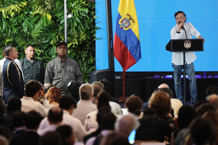 a man wearing a white shirt speaks at a podium on a stage, people seated in an audience face him