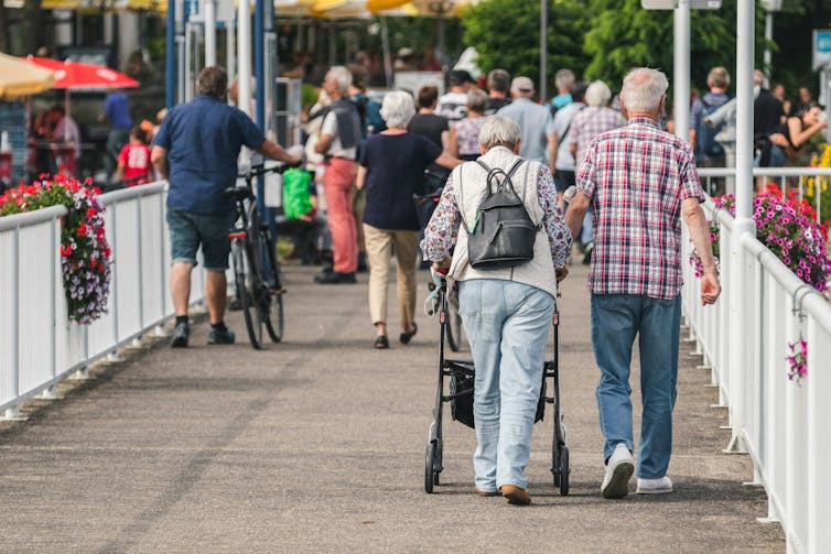 Elderly man walks arm-in-arm with an elderly woman using a walker.