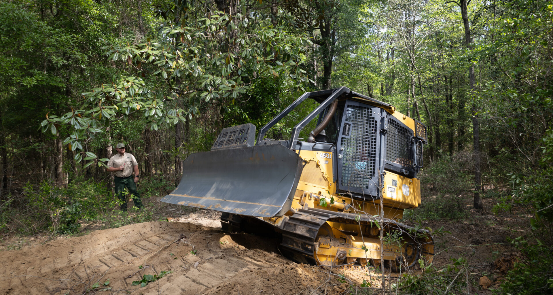 A yellow and black bulldozer climbs a small dirt hill in a forest of tall green trees, driving from the left of the frame to the right. Past the bulldozer, a man in a tan shirt and green pants stands in the trees, holding a radio.
