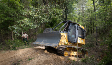 A yellow and black bulldozer climbs a small dirt hill in a forest of tall green trees, driving from the left of the frame to the right. Past the bulldozer, a man in a tan shirt and green pants stands in the trees, holding a radio.