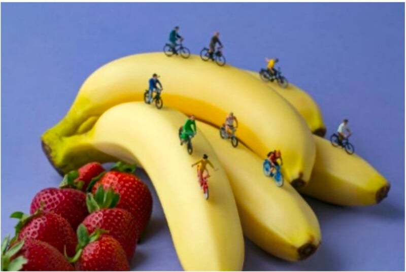 Miniature cyclists ride on a bunch of bananas with strawberries nearby, set against a purple background, creating a playful and imaginative scene.