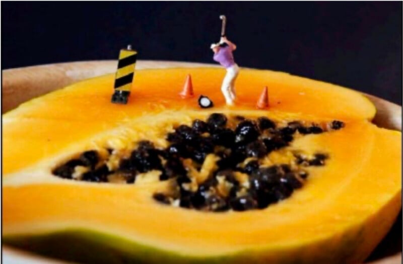 A miniature figure of a golfer is positioned on a sliced papaya, with two orange cones and a small striped barrier, making the fruit look like a whimsical golf course.