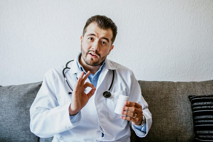 Male medical worker holding pill bottle and making okay gesture indoors on couch