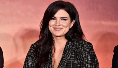 Gina Carano wears a v-neck blouse in front of a pink backdrop