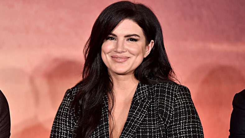 Gina Carano wears a v-neck blouse in front of a pink backdrop