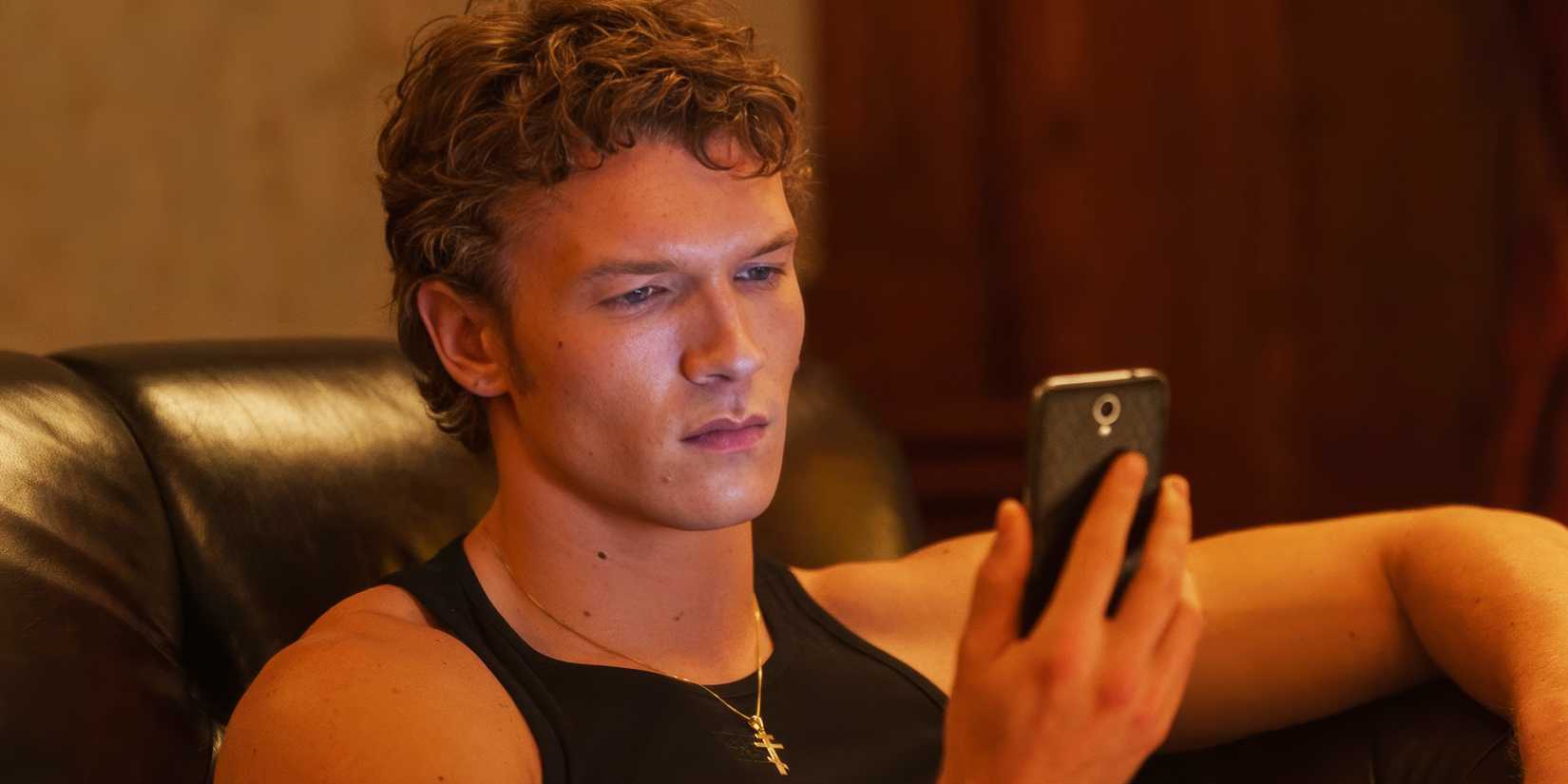 Connor Storrie as Ilya sitting on the couch and looking at his phone in Episode 5 of Heated Rivalry