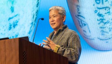 Hideo Mabuchi speaking on a lectern, with a photo of ceramics projected in the background