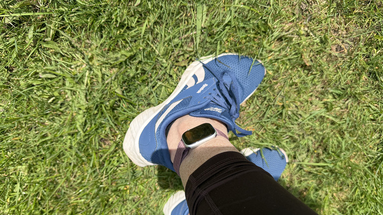 A side view of a woman's feet on grass with an Apple Watch around one ankle.