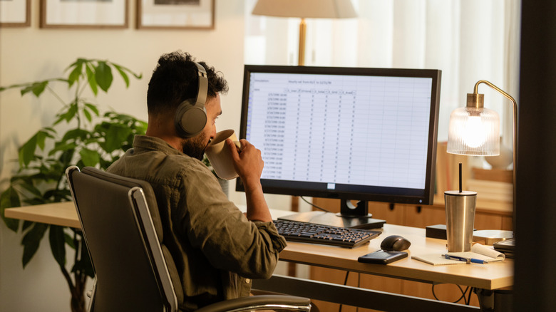 A man drinking a beverage while looking at a spreadsheet on a computer monitor.