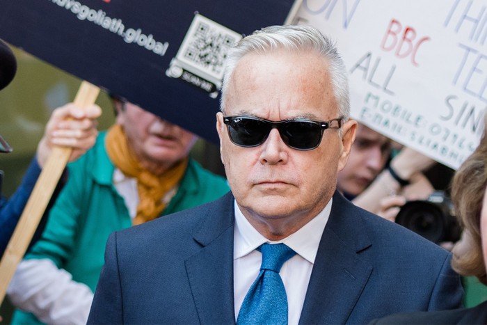 Huw Edwards wearing a blue suit and tie, with black sunglasses on looking ahead with a mute expression.