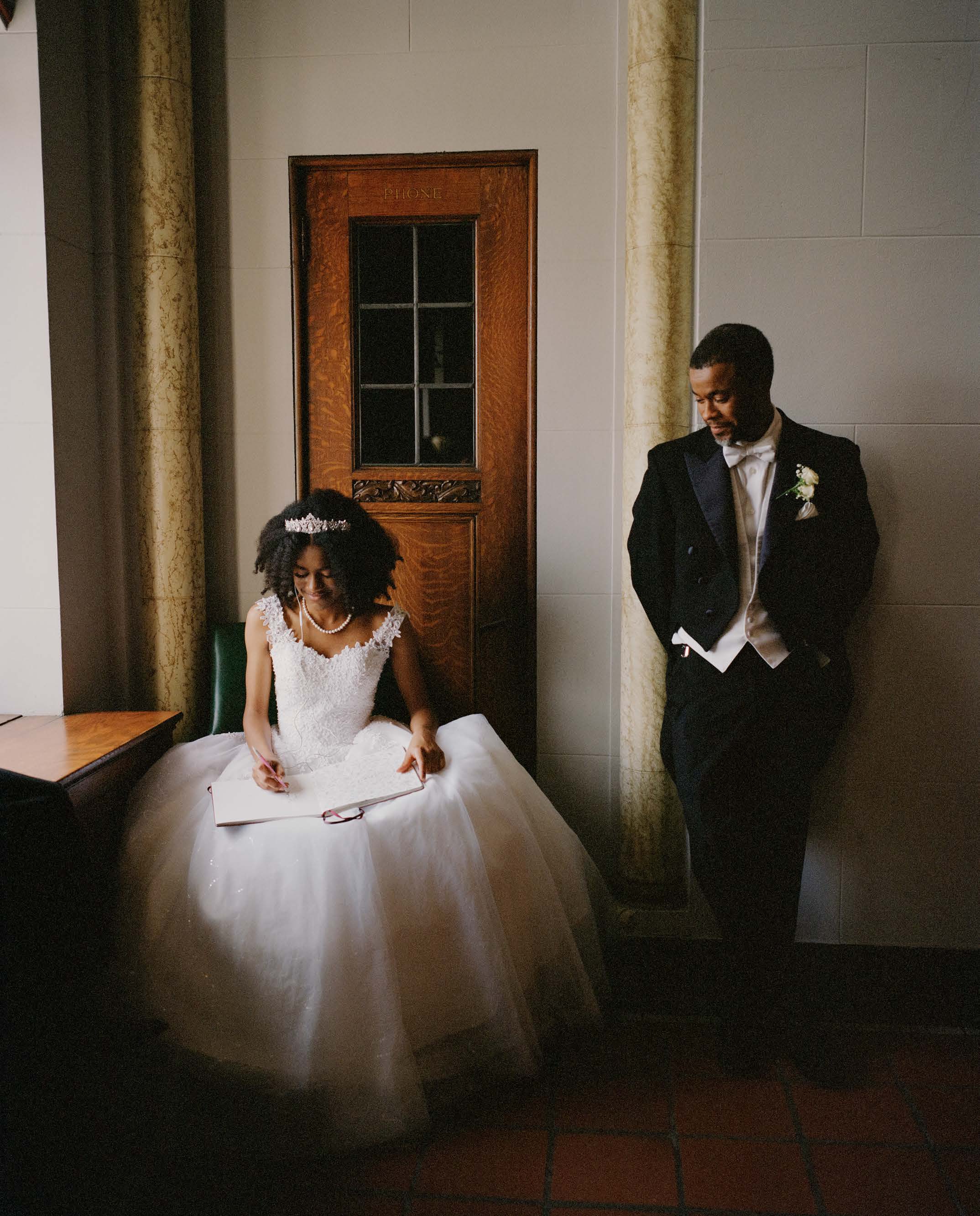 A young woman in a white ballgown and tiara writes in a book at a desk while a man in a tuxedo stands beside her, looking on, in a grand formal interior.