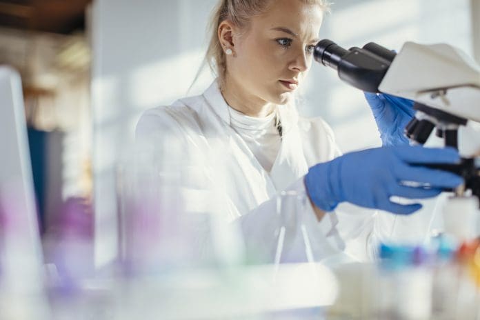 A Caucasian female lab technician attentively examines a specimen through a microscope, clad in professional lab attire, surrounded by scientific equipment in a bright, modern laboratory.