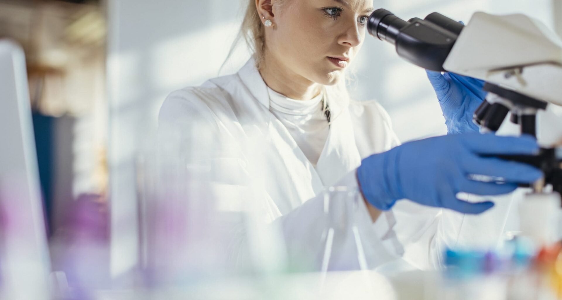 A Caucasian female lab technician attentively examines a specimen through a microscope, clad in professional lab attire, surrounded by scientific equipment in a bright, modern laboratory.
