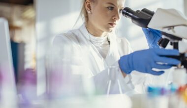 A Caucasian female lab technician attentively examines a specimen through a microscope, clad in professional lab attire, surrounded by scientific equipment in a bright, modern laboratory.