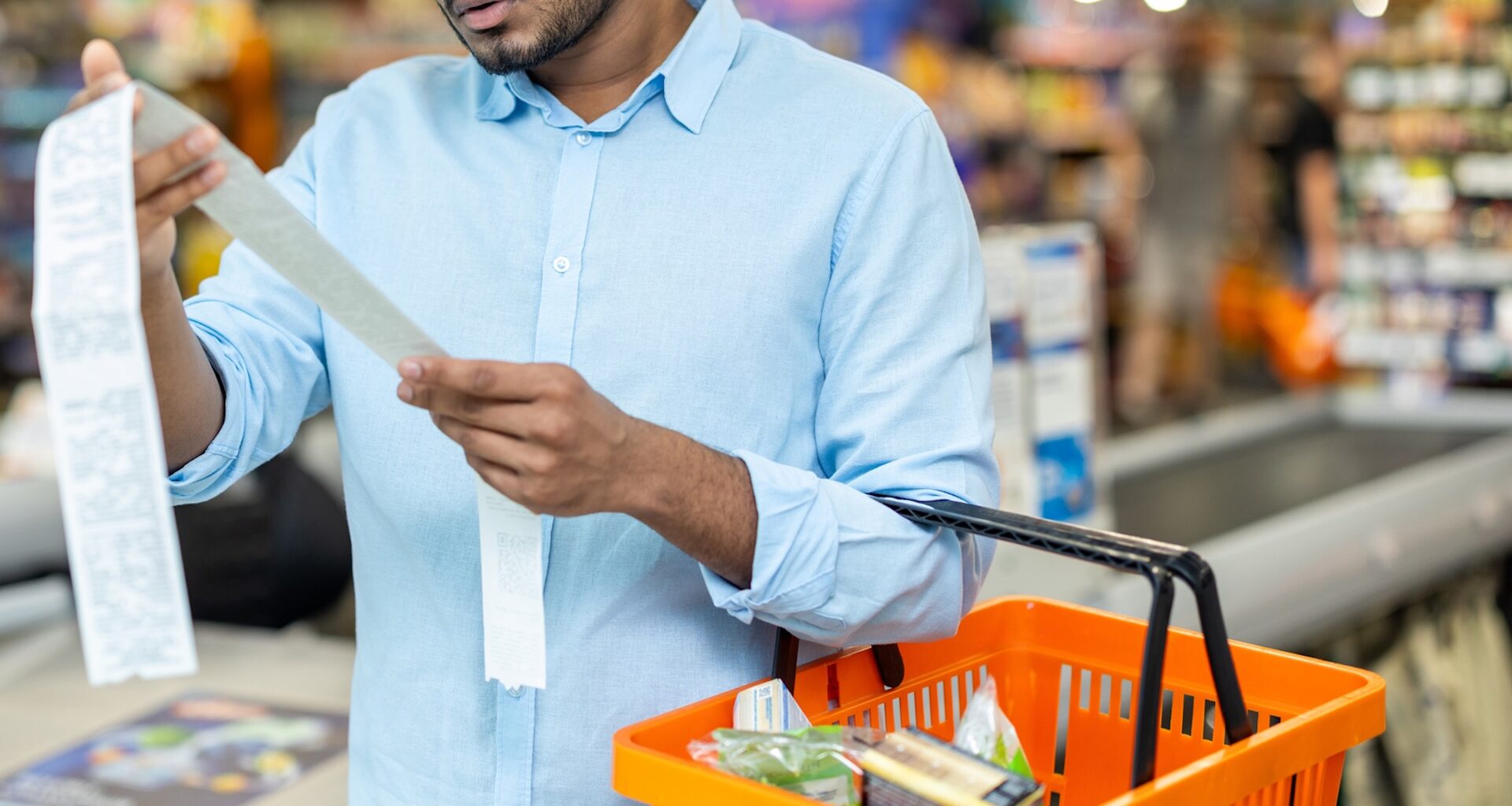 A man in a blue shirt examines a grocery receipt while holding an orange shopping basket in a supermarket.