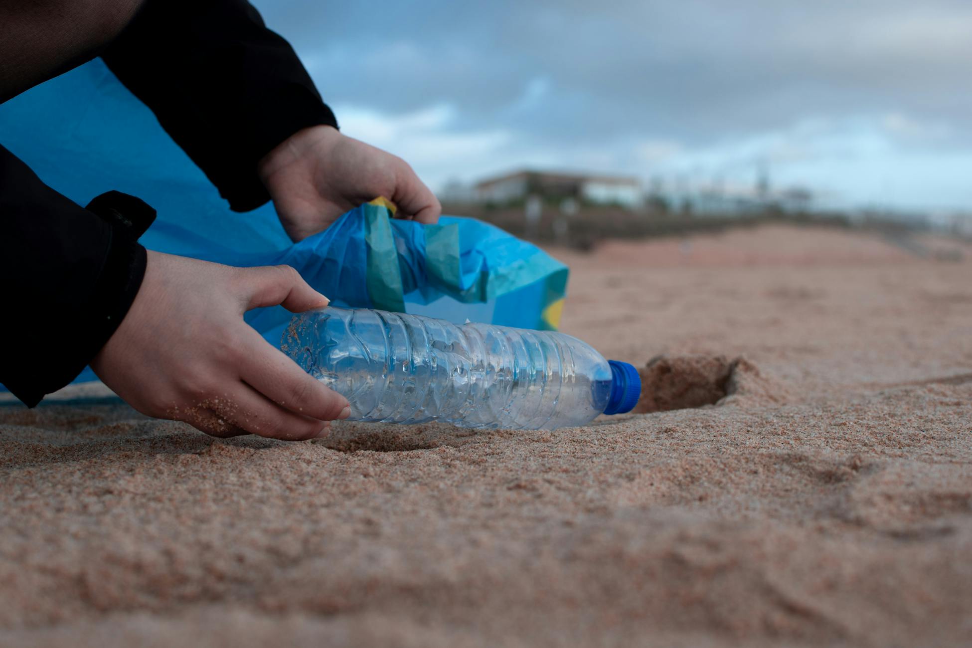 A hand collecting a plastic water bottle from a sandy beach.