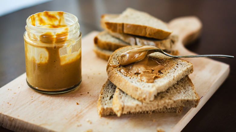 An opened, unnamed peanut butter jar is next to a spoon spreading peanut butter on the top piece of bread in a stack.