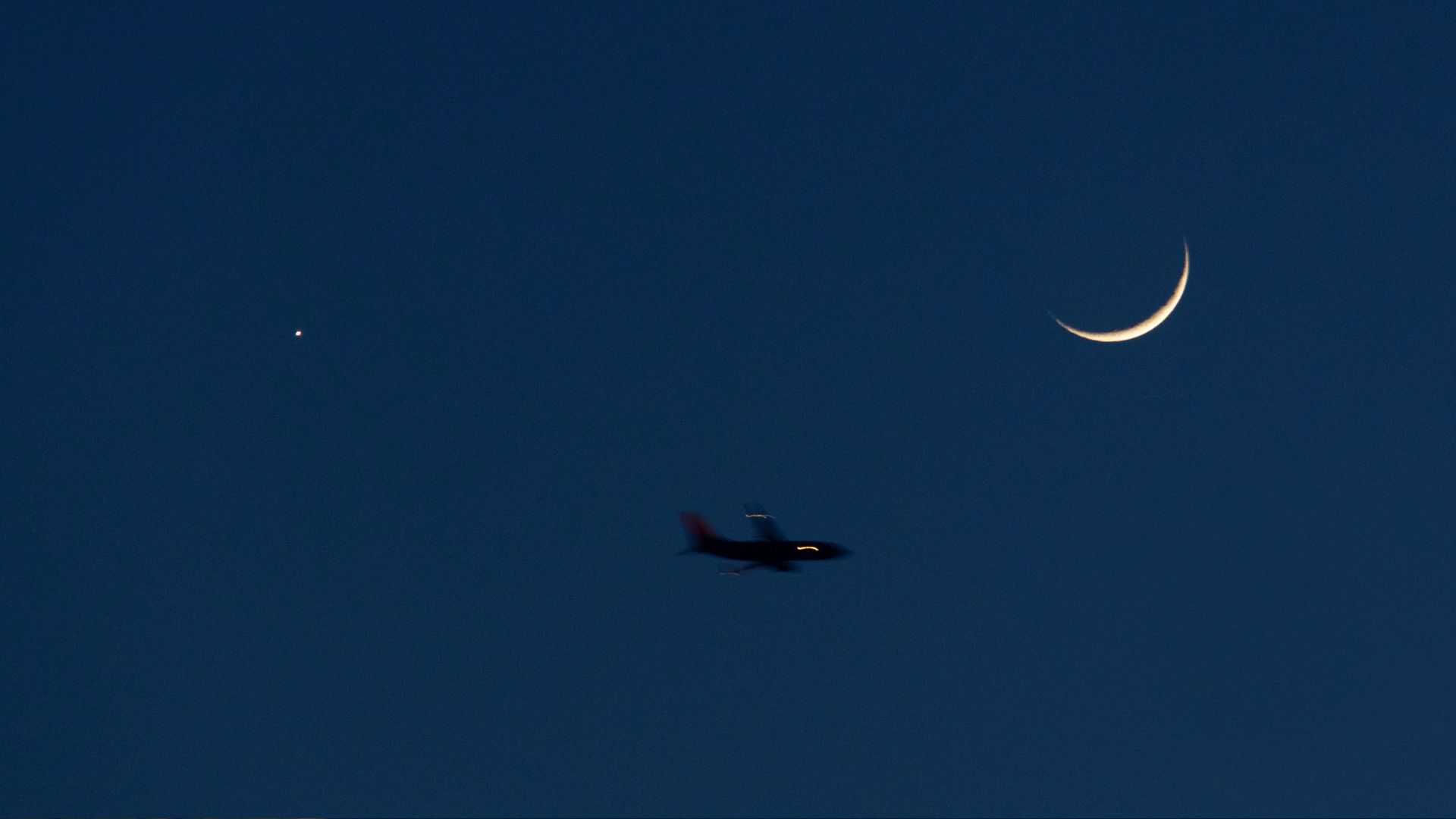 A plane is photographed flying below the crescent moon in a dark blue evening sky, as a star-like planet shines to its left.