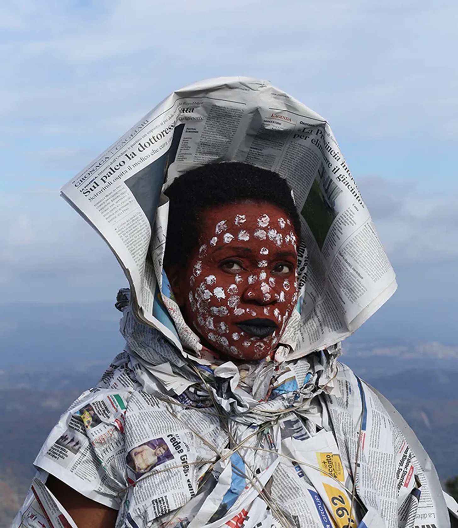 A person with face paint and black lips, wearing a costume and headpiece made from Italian-language newspapers, looks upward against a bright, cloudy sky.