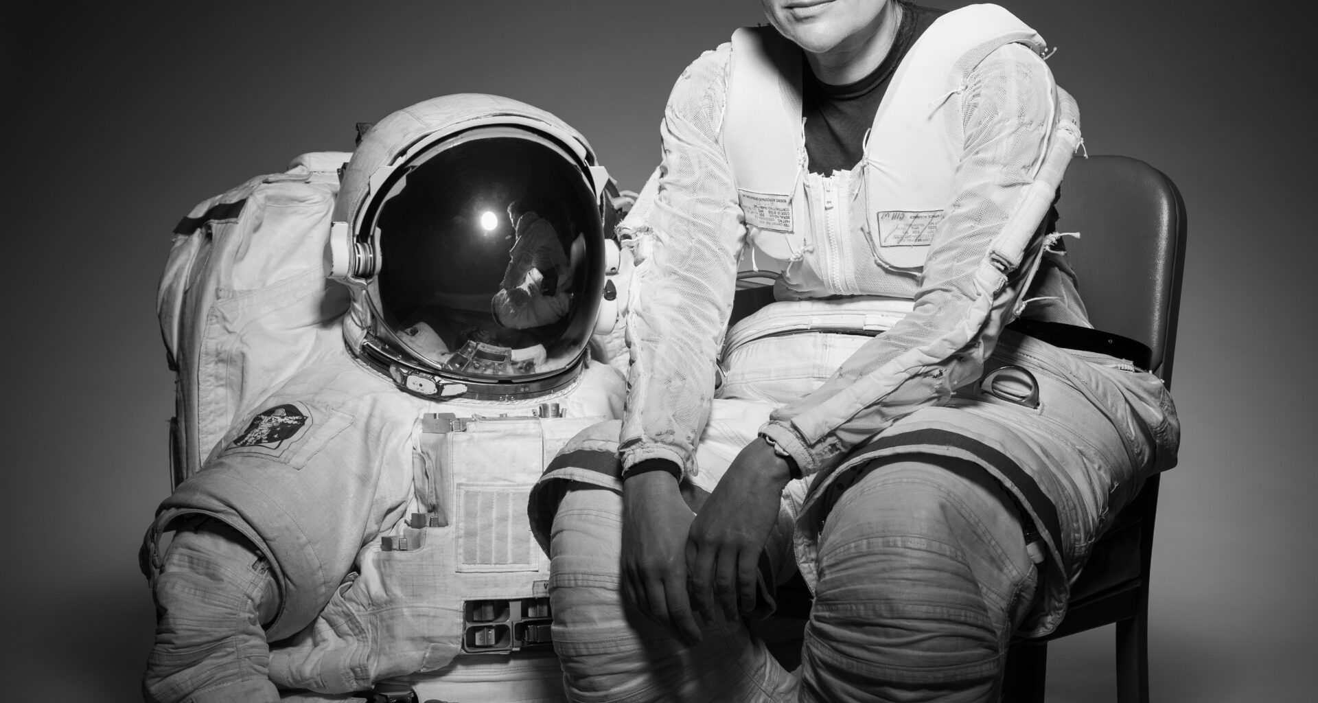 NASA Astronaut Jessica Meir sits in a chair. Her body is slightly turned to the right as she looks forward. She is wearing the bottom half of a spacesuit; the top half and gloves are on the floor next to the chair.