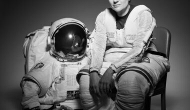 NASA Astronaut Jessica Meir sits in a chair. Her body is slightly turned to the right as she looks forward. She is wearing the bottom half of a spacesuit; the top half and gloves are on the floor next to the chair.