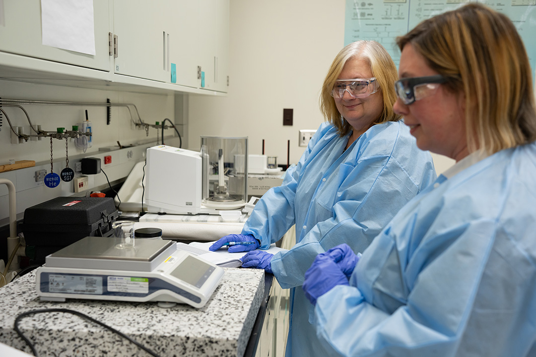 Two women in blue paper lab coats and safety glasses stand at a lab counter, looking at a small glass container on a scale.