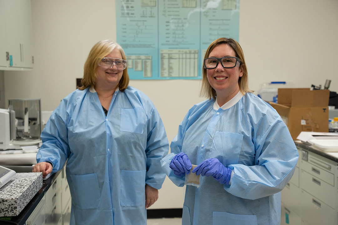 Two women in blue paper lab coats and safety glasses stand smiling in the lab.