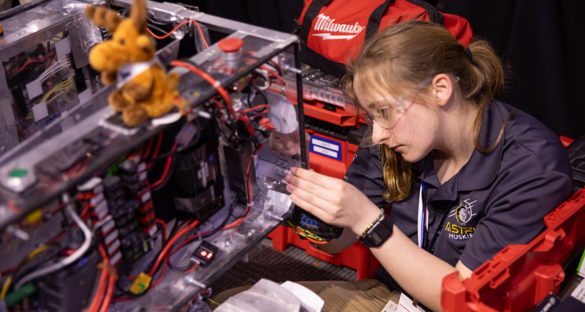 A student holds onto the edge of the rectangular body of a prototype lunar robot as she works on it. The rover has a rectangular body, which has multicolored attached to it. There is a small stuffed moose sitting on top of the rover.