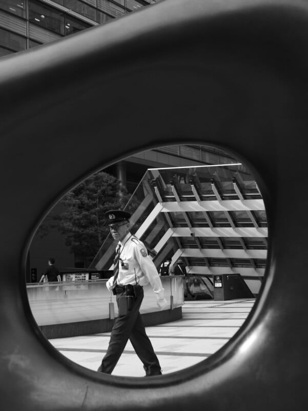 A security guard in uniform walks outside a modern building, seen through a large circular sculpture, creating a framed effect. The photo is in black and white.