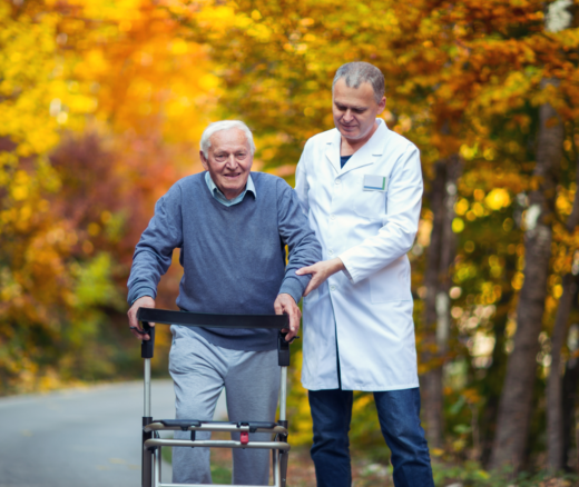 An older adult with a walker being led by a caretaker on a street with fall foliage.