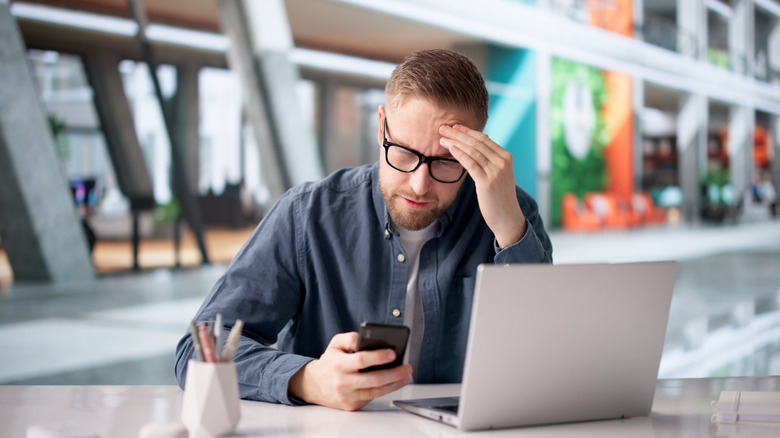 A man working at a computer and looking at his phone with a worried expression